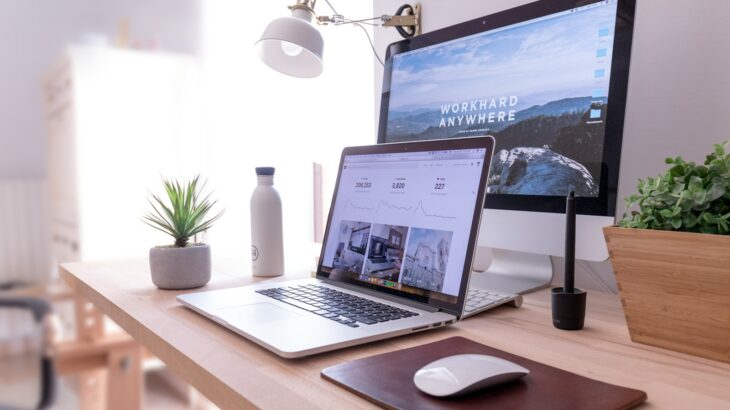 木製デスクに置かれたMacBook ProとiMac、マウス、観葉植物